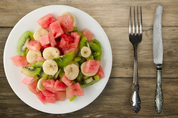 茶色の木の背景に健康的な新鮮なフルーツサラダの白いプレート フォークとナイフで果物サラダ — ストック写真
