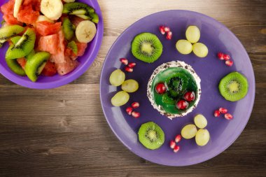 Kek ve meyve üzümleri, kivi, kahverengi ahşap zemin üzerinde nar. Mor tabak manzaralı meyveli tatlı.