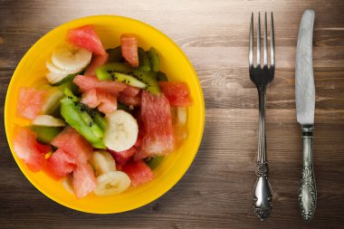Kahverengi ahşap tabakta sağlıklı taze meyve salatası. Çatal ve bıçak üst görüşlü vejetaryen salatası. .
