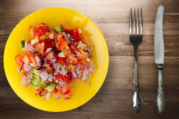 Vejetaryen yemeği. Biberli salata, domates, soğan, çatal ve bıçak manzaralı sarı tabakta brokoli. Kahverengi ahşap arka planda vegan salatası