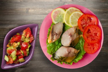 Pembe tabakta tütsülenmiş uskumru. Sebze salatanın üst manzarası. Siyah ahşap arka planda limonlu uskumru ve domates.