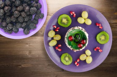 Kek ve meyve üzümleri, kivi, kahverengi ahşap zemin üzerinde nar. Mor tabak manzaralı meyveli tatlı.