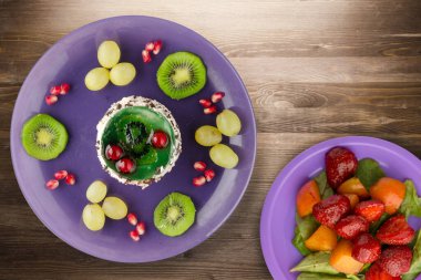 Kek ve meyve üzümleri, kivi, kahverengi ahşap zemin üzerinde nar. Mor tabak manzaralı meyveli tatlı.