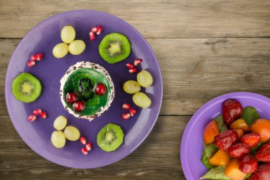 Kek ve meyve üzümleri, kivi, ahşap zemin üzerinde nar. Mor bir tabakta meyveli tatlı.