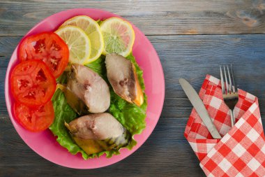 Çatal ve bıçak manzaralı pembe bir tabakta tütsülenmiş uskumru. Mavi ahşap arka planda limonlu uskumru ve domates.