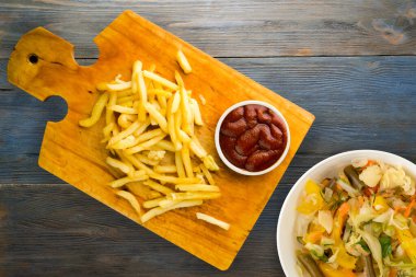 Gri mavi ahşap arka planda ketçaplı patates kızartması. Kahverengi ahşap tabakta patates kızartması ve vejetaryen salata.