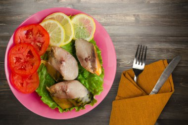 Çatal ve bıçak manzaralı pembe tabakta tütsülenmiş uskumru. Siyah ahşap arka planda limonlu uskumru ve domates.