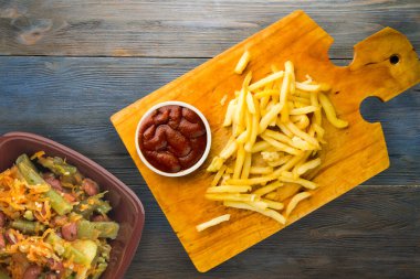 Gri mavi ahşap arka planda ketçaplı patates kızartması. Kahverengi ahşap tabakta patates kızartması ve vejetaryen salata.