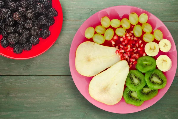 fruit mix pear, kiwi, grapes, banana, pomegranate on a green wooden background. fruit on a pink plate