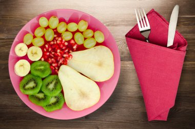 Meyve karışımı armut, kivi, üzüm, muz, kahverengi ahşap zemin üzerinde nar. Çatal ve bıçak manzaralı pembe bir tabakta meyve.