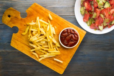 Gri mavi ahşap arka planda ketçaplı patates kızartması. Kahverengi ahşap tabakta patates kızartması ve vejetaryen salata.