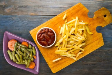 Gri mavi ahşap arka planda ketçaplı patates kızartması. Kahverengi ahşap tabakta patates kızartması ve vejetaryen salata.