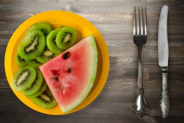 Sarı bir tabakta karpuz ve kivi dilimlenmiş. Ya da siyah ahşap bir zemin. Karpuz ve çatal ve bıçaklı kivi.