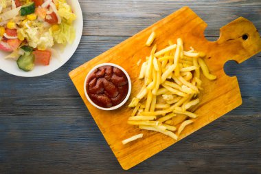 Gri mavi ahşap arka planda ketçaplı patates kızartması. Kahverengi ahşap tabakta patates kızartması ve vejetaryen salata.