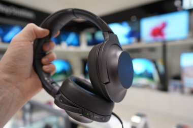 Gray headphones in a male hand in an electronics store. TVs in the background. clipart