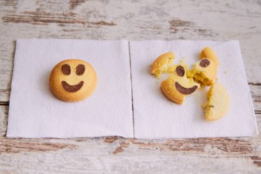 Gülümseyen emojiler şeklinde sarı kurabiyeler peçetenin üzerinde yatarken, bir başka kırık kurabiye de yakınlardaki bir peçetenin üzerinde, yakın plan, iyi ve kötü ruh hali, üzüntü ve neşe ile yan yana yatıyor..
