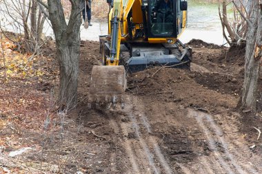 The excavator bucket reaches down to begin the first stage of soil extraction on a site with trees, excavator operation in construction during autumn.