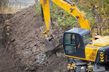 An excavator with a bucket full of soil in motion on a land plot.