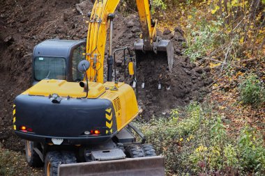 An excavator digging earth with its bucket at a construction site among trees and bushes, close-up of excavator performing earthworks, excavator dumping soil from the bucket.