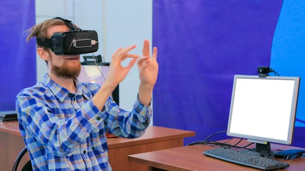 Young man using virtual reality headset in front of white blank monitor