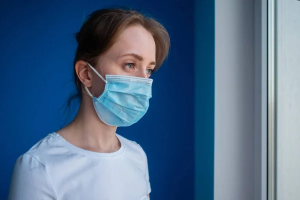 Pensive woman wearing medical face mask and looking out of window - side view