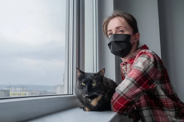 Woman in medical face mask and black cat looking out of window