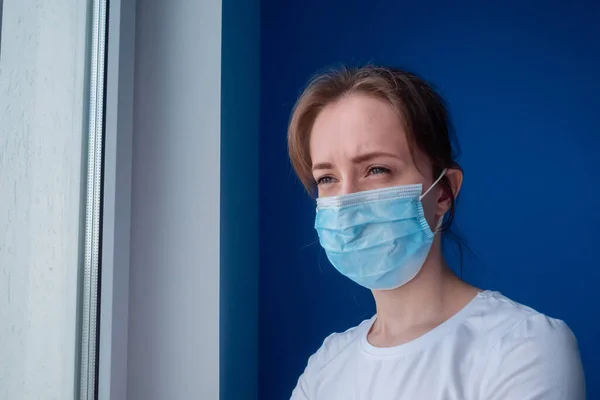 Pensive woman wearing medical face mask and looking out of window at home