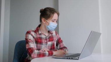 Yüz maskeli bir kadın dizüstü bilgisayar kullanıyor, görüntülü konuşma yapıyor - kişisel izolasyon kavramı