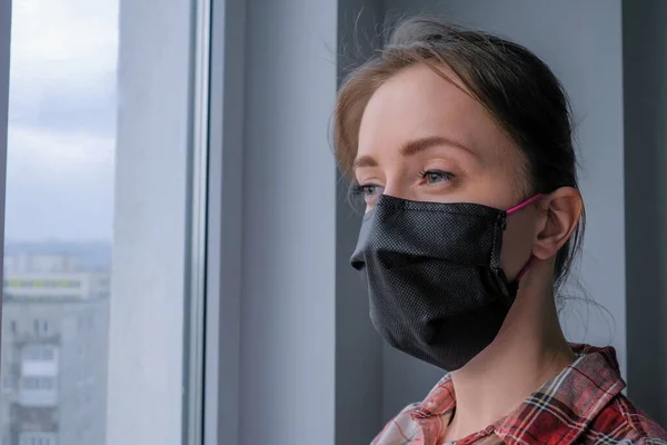 Pensive woman wearing medical face mask and looking out of window - close up