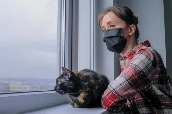 Black cat and woman in black medical face mask looking out of window at home