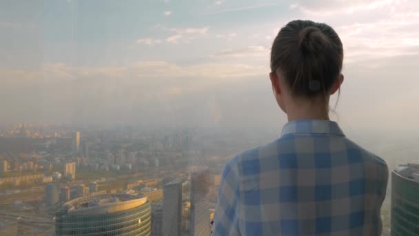 Задний вид женщины, смотрящей на захватывающий городской пейзаж через окно небоскреба — стоковое видео