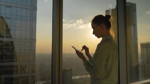 Mujer usando teléfono inteligente contra la puesta del sol vista del paisaje urbano a través de la ventana — Vídeos de Stock