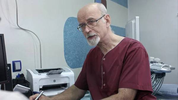 Зрілий лікар за своїм столом, зробити кілька нотаток, слухаючи старшого пацієнта — стокове фото