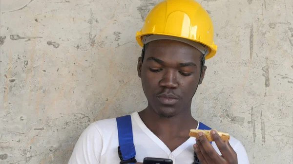 Hungry afro-american worker at construction site eating sandwich lunch food looking at smartphone