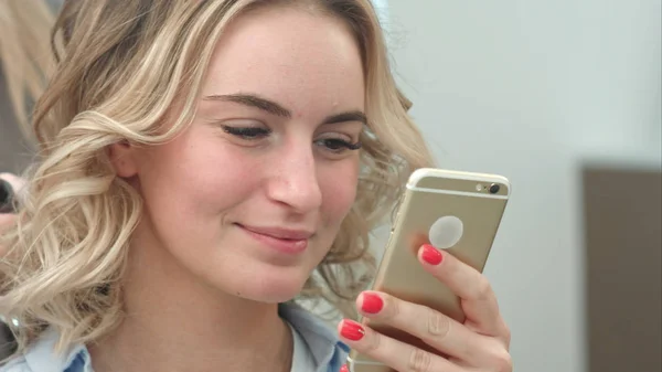 Hermosa mujer usando teléfono inteligente en un salón de belleza, mientras que tener un peinado — Foto de Stock