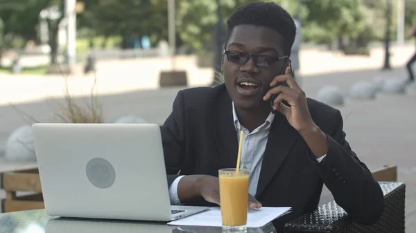 Young attractive afro american businessman with glasses and laptop sitting in cafe bar and talking on cell phone