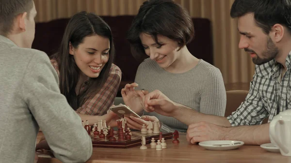 Группа молодых людей, играющих в шахматы и использующих смартфон — стоковое фото