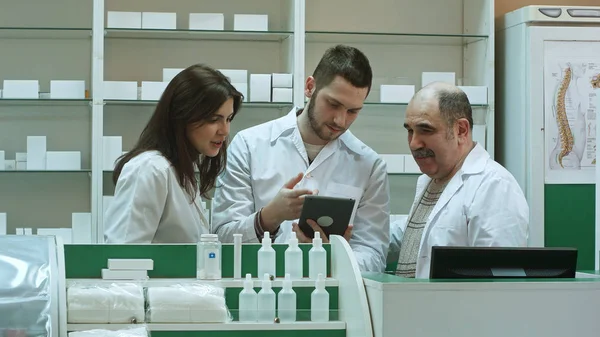 Equipe sorridente de farmacêuticos, dois homens e uma mulher, ficam lado a lado na farmácia verificando informações em um computador tablet — Fotografia de Stock