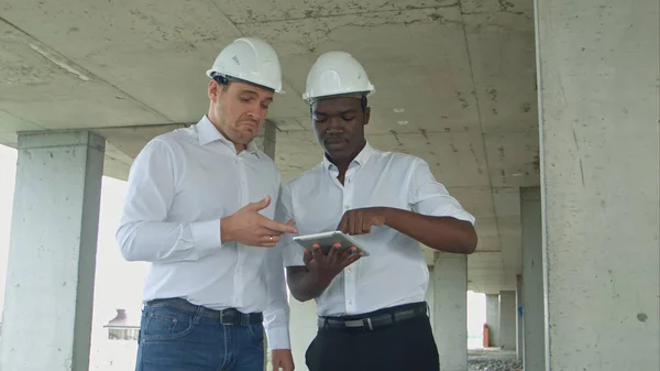 Engenheiro afro-americano e arquiteto caucasino usando tablet digital e usando capacetes de segurança no canteiro de obras — Fotografia de Stock