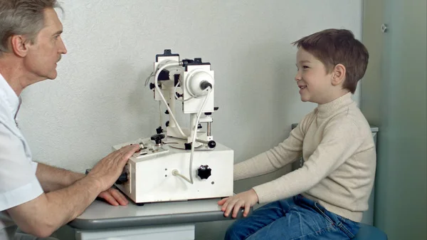 Okulista w pokoju egzamin z mały chłopiec siedzący w fotelu patrząc na oko test maszyna — Zdjęcie stockowe