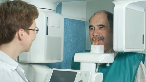 Врач-стоматолог разговаривает со старшим пациентом перед обследованием с помощью панорамного и цефалометрического рентгеновского сканера — стоковое фото