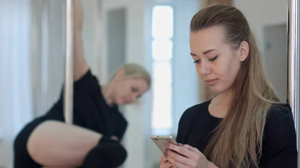 Dançarina de pólo bonito fazendo uma pausa de seu treino e redes sociais com um telefone celular, enquanto seu parceiro fazendo exercícios — Fotografia de Stock