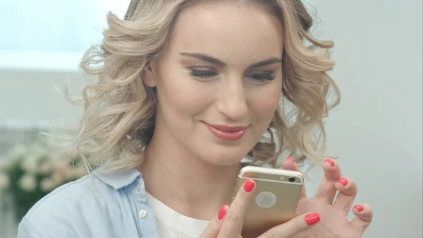 Chica rubia está sentada en una sala de estar y mirando la pantalla del teléfono inteligente sonriendo — Foto de Stock