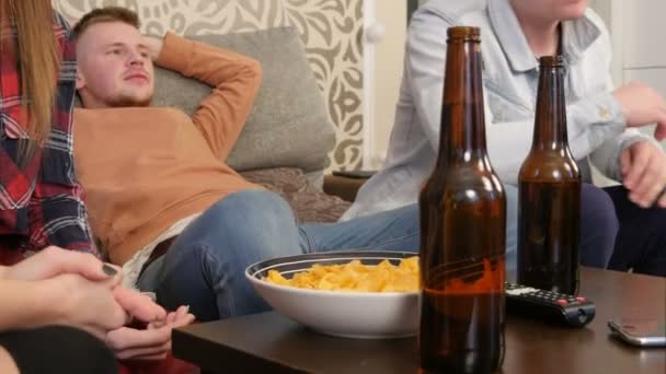 Группа друзей, сидящих на диване, вместе смотрящих телевизор и пьющих пиво — стоковое видео