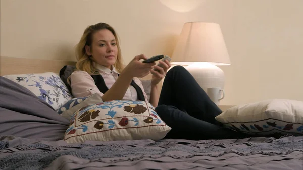 Jovem mulher bonita deitada na cama, ligando os canais de TV e usando o smartphone em casa — Fotografia de Stock