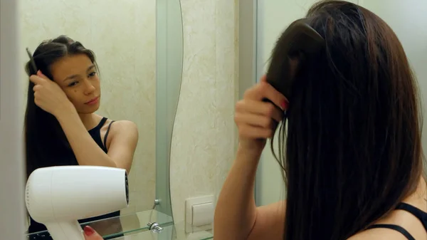 Hermosa chica morena secándose el pelo y mirándose en el espejo en su baño — Foto de Stock
