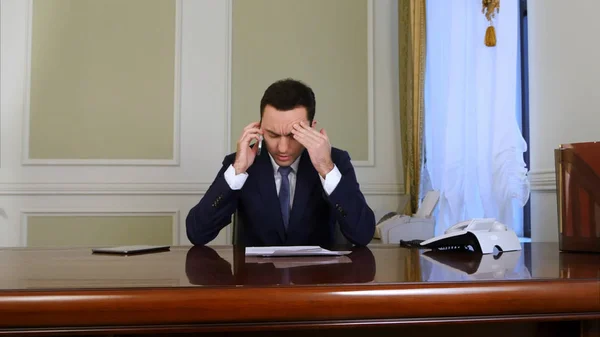 Podnikatel hledá unavený a mluví o mobilní telefon při sezení v kanceláři — Stock fotografie