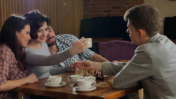 Молодая пара играет в шахматы в помещении с друзьями и делает селфи — стоковое фото