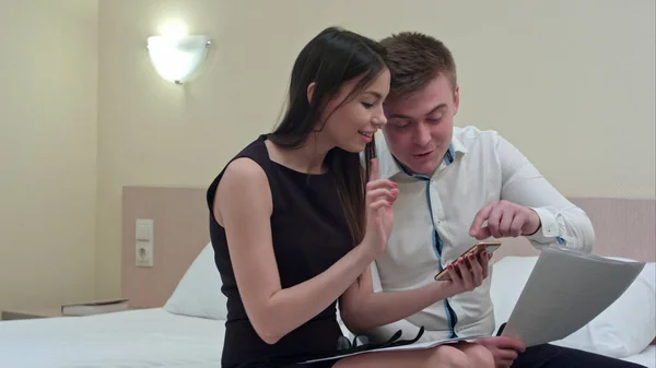 Belo casal sentado na cama e usando smartphone, parecendo felizes juntos — Fotografia de Stock