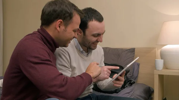 Молодые друзья-мужчины, сидящие на диване и пользующиеся планшетом, что-то осуждающие и смеющиеся — стоковое фото